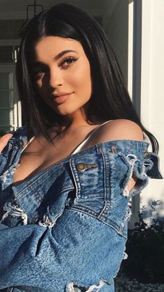 a woman with long black hair wearing a jean jacket and ripped jeans standing in front of a house