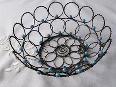 a metal basket with blue beads and black wire on a white tablecloth covered surface