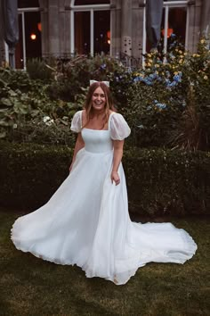 a woman in a white wedding dress standing on the grass with her arms behind her back