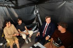 four people are sitting in a tent with one man looking at the camera and two women standing behind him