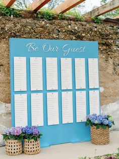 a blue and white wedding seating chart with flowers in baskets next to it on the side of a stone wall