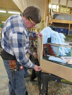 a man in plaid shirt working on a model train at a table with other items