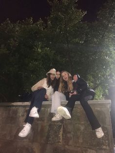 three young women sitting on the edge of a stone wall at night with trees in the background