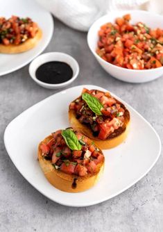 three small sandwiches with tomatoes and basil on top, sitting on a white plate next to some dipping sauce