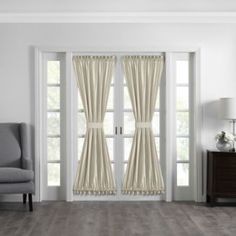 a living room with white walls and wooden flooring, two windows covered in beige drapes