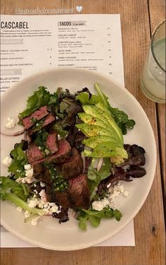 a steak salad with lettuce and feta cheese on a white plate next to a glass of water