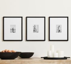 three black and white photographs hang on the wall above a bowl of eggs in front of some candles