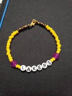 Simple yellow beaded bracelet with purple accent beads and it reads "Lakers" This is unisex adult standard size Casual Purple Friendship Bracelets With Letter Beads, Casual Purple Friendship Bracelet With Letter Beads, Customized Purple Beaded Friendship Bracelets, Adjustable Yellow Friendship Bracelets With Letter Beads, Personalized Adjustable Yellow Friendship Bracelets, Yellow Letter Beads Friendship Bracelets As Gift, Personalized Adjustable Yellow Beaded Bracelets, Casual Yellow Friendship Bracelets With Letter Beads, Gift Yellow Letter Beads Friendship Bracelets