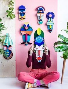 a woman sitting on the floor holding up a mask in front of her face, surrounded by plants