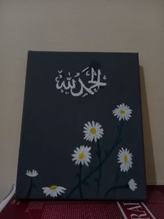 a painting with white daisies and arabic writing on the bottom half of it, against a beige wall