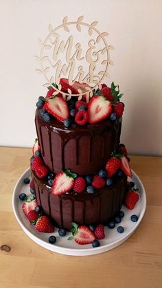 a three layer chocolate cake with strawberries and blueberries on the bottom, topped with a wooden monogram