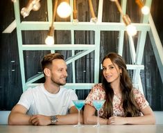 a man and woman sitting at a table with drinks in front of them, posing for the camera