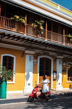 a woman riding a red scooter down a street next to a tall yellow building