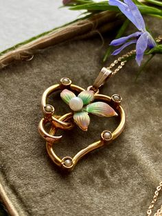 A 9CT gold Art Nouveau Style enamel floral heart pendant set with a border of seed pearls.:The hand painted enamel flower is in subtle shades of green and pink. The flower has a central Pearl. The scrolling border has all the whiplash  features of the Art Nouveau movement and softened with the addition of five small seed pearls. The piece has some age as the seed Pearl colour has darkened with age.  The bail is a simple wedge shaped one and the fine trace chain is modern and 9CT gold. The price Vintage Flower Necklaces For Anniversary, Antique Enamel Necklaces For Wedding, Antique Enamel Necklace For Wedding, Art Nouveau Gold Enamel Jewelry, Gold Enamel Art Nouveau Jewelry, Vintage Heart Enamel Necklaces, Vintage Enamel Heart Necklaces, Vintage Heart Enamel Necklace, Vintage Enamel Heart Necklace