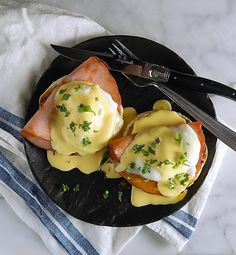 an open face sandwich with hollandaise sauce on a black plate next to a knife and fork