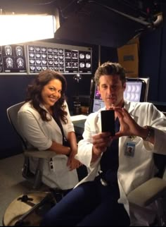 a man and woman sitting next to each other in front of a computer desk holding up a cell phone