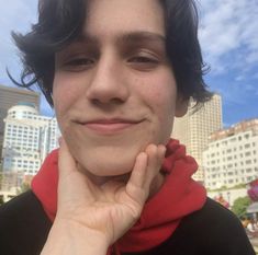 a young man with black hair and a red scarf smiles at the camera in front of a cityscape