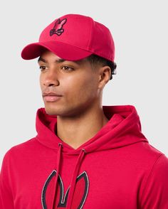 A person wearing the Psycho Bunny MENS WALTER BASEBALL CAP - B6A800D200 and a matching red hoodie with a bunny logo, looking to the side against a plain background. The durable and comfortable cap is crafted from 100% cotton, perfectly complementing the casual outfit. Cold Weather Hats, Bunny Man, Bunny Logo, Polo Tees, Swimwear Shorts, Active Wear Outfits, Cotton Polo, T-shirt Polos, Sweater Jacket