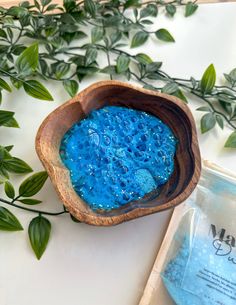 a wooden bowl filled with blue powder next to some green leaves on a white surface