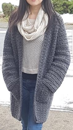 a woman standing in front of a body of water wearing a gray sweater and scarf