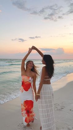 two beautiful women standing on top of a sandy beach next to the ocean at sunset