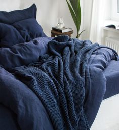 an unmade bed with blue sheets and blankets on it, next to a potted plant