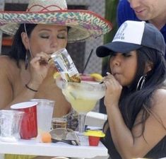 two women sitting at a table with drinks and hats on, one drinking from a straw