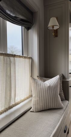 a window seat with two pillows and a lamp on the wall next to it in front of a window