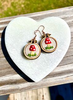 a pair of earrings that are on top of a heart shaped piece of wood with grass and mushrooms in it