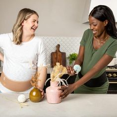 two women in the kitchen preparing food and laughing at each other's company while one woman holds a vase