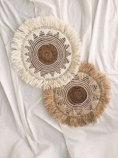 two woven baskets sitting on top of a white bed next to each other, one with fringes