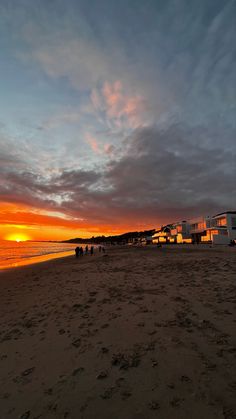 people are walking on the beach as the sun sets in the distance with buildings behind them