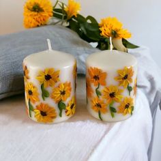 two white candles with yellow flowers on them sitting on a bed next to a pillow