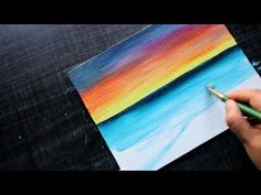 someone is holding a paintbrush and painting a colorful sunset on a piece of paper