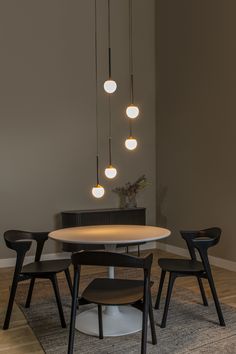 a dining room table and chairs with lights hanging from the ceiling above it, in front of a gray wall