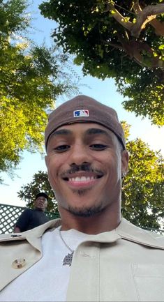 a man wearing a baseball cap smiles at the camera while standing in front of trees