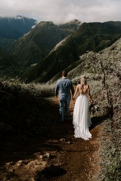 a bride and groom walking down a dirt path in the mountains on their wedding day