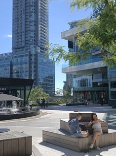 two people are sitting on benches in the middle of a plaza with tall buildings behind them