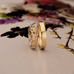 two gold wedding rings sitting on top of a floral print table cloth with flowers in the background