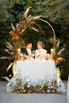 a couple sitting at a table with candles in front of them and surrounded by greenery