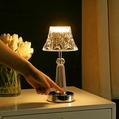 a person reaching for a flower in a glass vase on a table next to a lamp