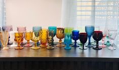 many different colored wine glasses lined up on a table next to a window with sheer curtains