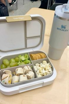 an open lunch box with grapes, crackers and cheese in it next to a coffee cup