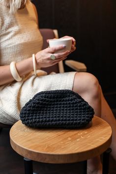 a woman sitting on a stool holding a cup and a black crocheted purse