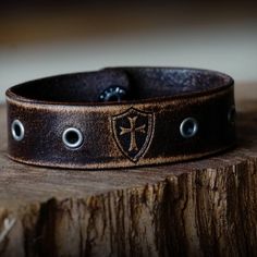 Made from premium grade 7 ounce leather. Comes engraved with the Crusader cross and shield shown centered between gunmetal eyelets. Strap is .75 inches wide. Hand dyed, distressed, oiled and finished in house. Made in America. Please note that adding personalization will replace the engraving shown. Instructions can be added in the note at checkout. Rustic Leather Concho Bracelets, Vintage Leather Concho Bracelets, Vintage Leather Bracelet With Engraving, Brown Leather Concho Bracelet, Vintage Engraved Leather Bracelets, Vintage Leather Engraved Bracelet, Vintage Leather Jewelry With Waxed Finish, Vintage Distressed Brown Leather Bracelet, Adjustable Leather Gothic Wristband