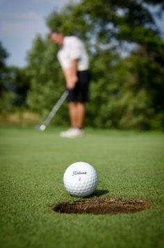 a golf ball sitting on top of a green field next to a hole with a man in the background