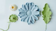 three crocheted flowers and two leaves on a white surface, one is blue and the other is green