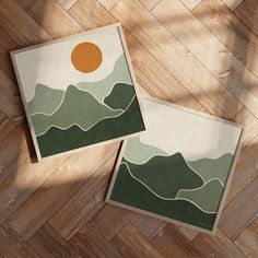 two framed pictures sitting on top of a wooden floor next to each other with mountains in the background