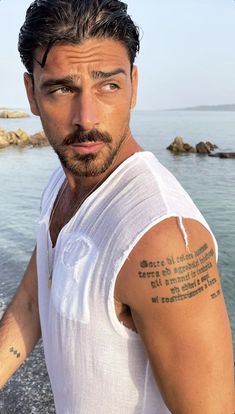 a man with a tattoo on his arm standing next to the ocean and looking at the camera