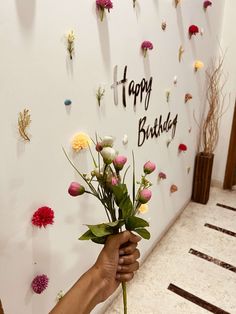 a person holding flowers in front of a happy birthday sign on a wall with paper flowers